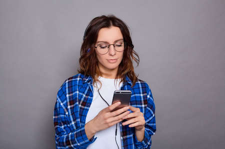 Caucasian young beautiful serious woman posing in blue shirt and white t-shirt isolated over gray wall background listening music with headphones using smartphone.の写真素材