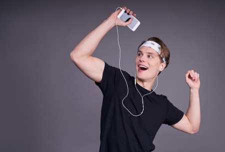 A young white man dances, makes movements to music, has modern stereo headphones, smiles positively, being in high spirits. Wearing a black t-shirt and white bandana.の写真素材