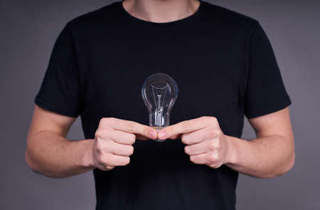Close-up, a young white guy in a black base T-shirt holds a light bulb with two hands. The concept of energy saving, resources, electricity.の写真素材