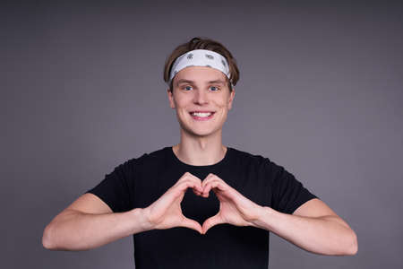 Charming handsome young man in a basic black T-shirt, makes his hands heart, in love and in a good mood, squished against a gray background.の写真素材