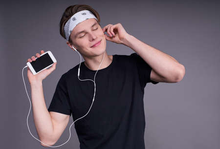 A calm, handsome man, a student, in a black basic T-shirt, listens to music with headphones through his smartphone, squished against a grey background. A copy of the space.の写真素材