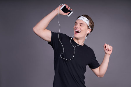 The cheerful joyful young guy is dancing, making movements to music, has modern stereo headphones,...