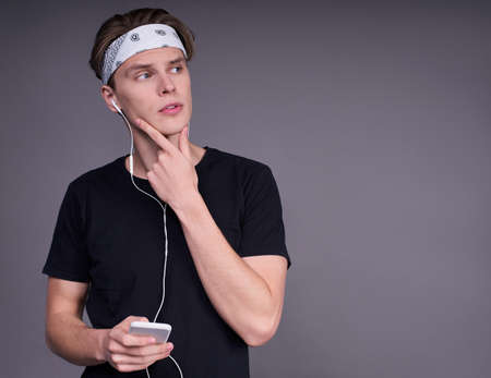 Pensive young Caucasian guy, a student in a black T-shirt, wears headphones in his ears, holds the phone in his hand, touches his chin with his hand, looks to the right, stands on a gray background.の写真素材