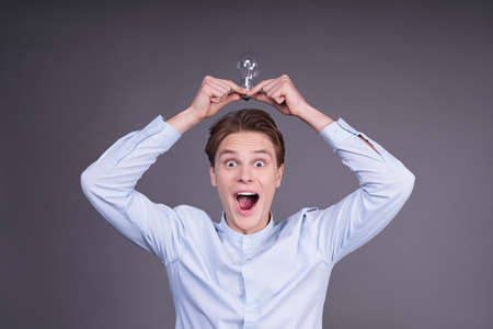 Good idea! Handsome young excited cheerful glad, delightful handsome shocked astonished surprised man in a blue shirt with a light bulb over his head, isolated on gray background.の写真素材