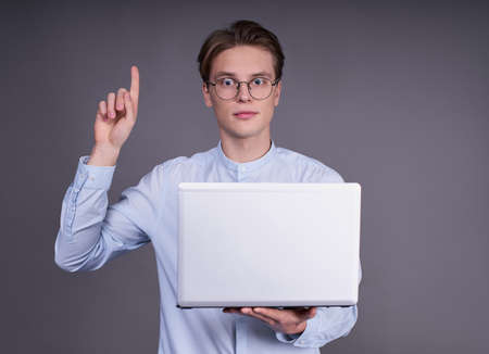 Charming calm young handsome guy, student, businessman, manager in a blue shirt and glasses, holds a silver laptop in his hands, raised his finger up, has a good idea, stands on a gray background.の写真素材