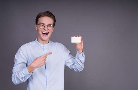 Cheerful happy young white guy in glasses and a blue shirt, holds a white card in his left hand and points at it with his finger, looks directly, stands on a gray background.の写真素材