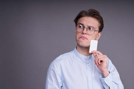 Smiling guy choosing between online purchases. A young Caucasian man in a blue shirt, holds a credit card in his hand, is thinking of making an online purchase. Finance, money, advertising concept.の写真素材