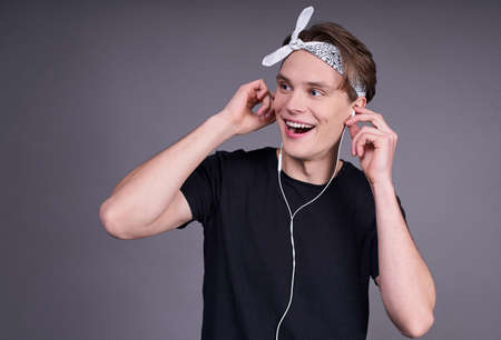 Happy joyful cheerful cute guy, student, in a black basic T-shirt, listening to music with headphones, looking right, squished against a gray background. A copy of the space.の写真素材