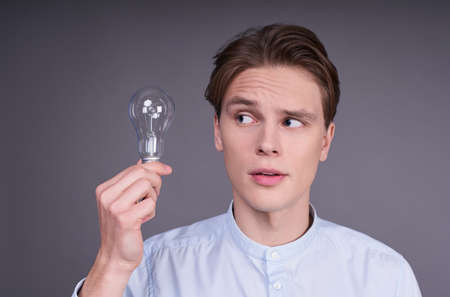 Close up portrait of an emotional smart cheerful student in a blue shirt holding a light bulb and founded a great new solution, standing on gray background.の写真素材
