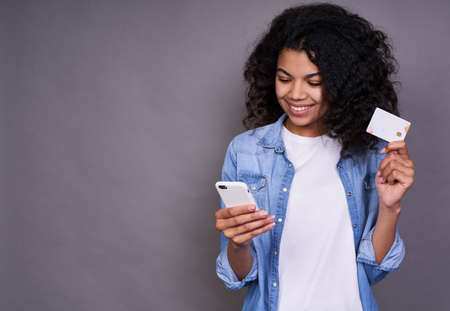 Portrait of a happy young African, American woman in a white T-shirt and denim shirt, showing a plastic credit card while using a mobile phone isolated on a gray background. Online shopping concept.の写真素材