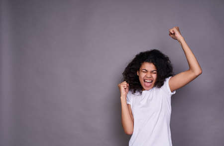 Cute charming young African woman, in a white basic T-shirt, having fun, dancing, rejoices, raised her hands up, posing on a gray background.の写真素材