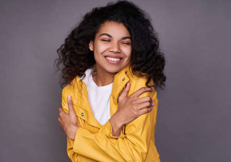 Young tender vulnerable young beautiful dark-skinned girl with black curly hair, in a white T-shirt and yellow jacket, hugs herself with her arms, dreamily closes her eyes and smiles.の写真素材