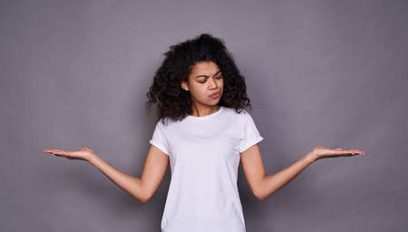 A young cute cute African woman, about 20 years old, in a white basic T-shirt, standing on a gray background, making a choice, spread her arms to the side, looking to the side.の写真素材