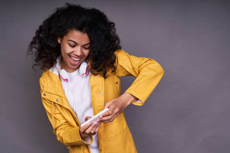 Cute cheerful joyful playful young African girl, in a white T-shirt, with magnificent curly hair, holds a smartphone in her hand, plays online games, a young gamer.の写真素材
