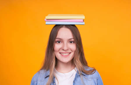 Young blond Caucasian girl with books on her head close-up, looks cute and gentle, smiles with a broad smile, looks right on a yellow background.の写真素材