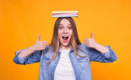 Young cute blond Caucasian girl with books on her head, looks happy and joyful, smiles broadly, showing excellent with both hands, ok, like, looks right on a yellow background.の写真素材