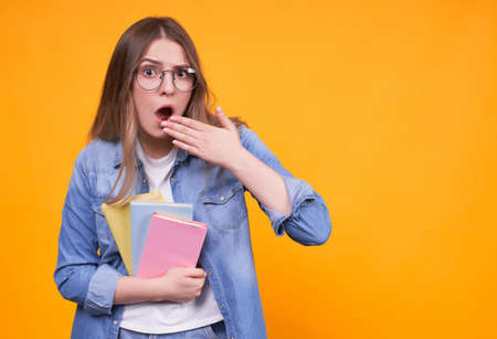 Young Caucasian surprised funny girl in glasses with notebooks, looking surprised and unexpected, wants to cover her open mouth, thinking or making choices on a yellow background, copy space.の写真素材