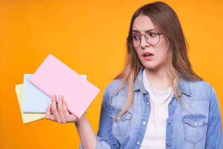 Emotional freckled woman covers her mouth, scared, has a notebook, wears a casual white t-shirt and jeans, in glasses, posing on a yellow background. The concept of people, study and emotions.の写真素材