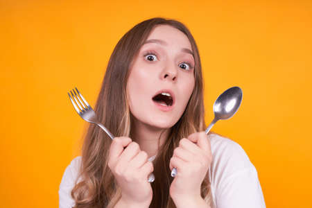 Frightened and unhappy young Caucasian girl with open mouth, bewildered, crossed a fork and a spoon in her hands, on a yellow background.の写真素材