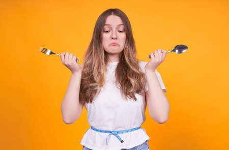 Unhappy young Caucasian girl with a fork and a spoon in her hands, dissatisfied with her weight, appearance, a woman with a tape measure on a yellow background.の写真素材