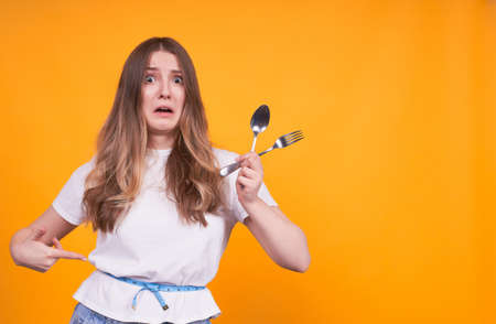 Unhappy young Caucasian girl with a fork and a spoon in her right hand, and her left indicates her waist, dissatisfied with her weight, appearance, a woman with a tape measure.Cope space.の写真素材