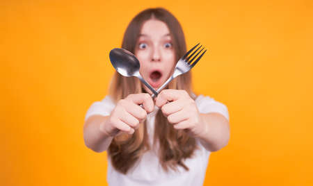 A frightened and unhappy young Caucasian girl crossed her fork and spoon in her hands, the focus is on the spoon and fork, dissatisfied with her weight, appearance, on a yellow background.の写真素材