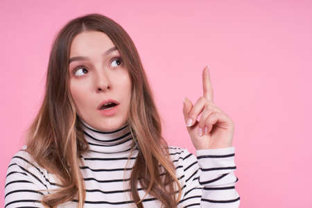 Emotional young pensive blonde lady looks up right, close-up, feels something impressed, points with her index finger, wears a casual striped sweater isolated on a pink background, copy space.の写真素材