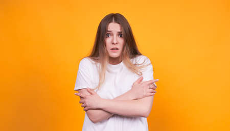 A young, pretty, surprised Caucasian girl, in a white basic T-shirt, hugged herself with her arms, with wide open eyes, looks straight. Photo on a yellow background.の写真素材