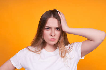 Young nice blonde girl with long hair, in a white basic t-shirt, touches her head with her hand. Headache, irritation, fatigue. Posing on a yellow background.の写真素材