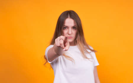 Unhappy caucasian woman in white t-shirt, pointing finger at camera isolated on an orange background in studio. People sincere emotions, lifestyle concept.の写真素材