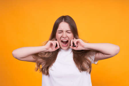 Young cute and pretty girl in a white T-shirt on a yellow isolated background, screaming and covering her ears with her hands, panicking, hysterical.の写真素材