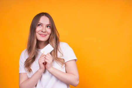 Young cute graceful caucasian girl holding a credit card in hands, over isolated yellow background, thoughtfully, cute smile, think what to buy,anticipation.Copy space.の写真素材