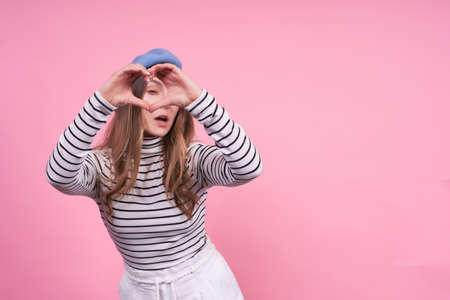 Young cute lady in a striped sweater, fashionable blue beret, full of love and enthusiasm, surprised with an open mouth, shows with two hands a heart sign, on a pink background, copy space.の写真素材