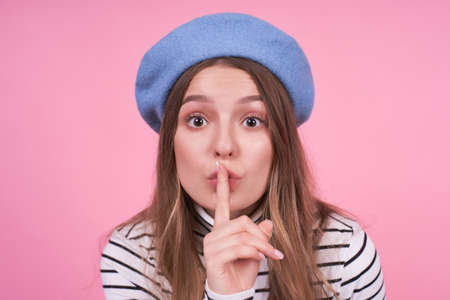 Close up portrait of a cute lovely attractive girl in a fashionable hat beret gesturing silence sign with index finger pout lips.to ask for silence or not to tell anyone your secret, raising eyebrows.の写真素材
