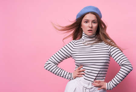 Portrait of a cute emotional passionate Caucasian girl, in a stylish fashionable beret hat with a calm face, holding hands on her hips, the wind develops long blond hair, striped sweater copy space.の写真素材