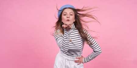 Portrait of a cute passionate emotional Caucasian girl in a stylish fashionable beret hat with a calm face, blows a kiss, the wind develops long blond hair, in a base striped sweater copy space.の写真素材