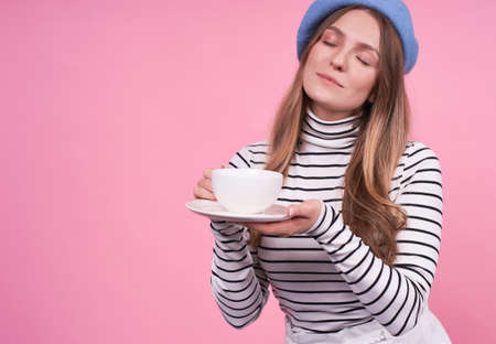 Portrait of a young cute Caucasian woman with a gentle, satisfied expression, in a blue stylish beret, holds tea, coffee, drink in a cup on a plate, copy space.の写真素材