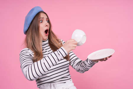 Portrait of a young cute Caucasian woman with a funny, surprised, scared face and open mouth, in a blue stylish beret, pouring tea, coffee, a drink in a cup and a plate in her right hand.copy place.の写真素材