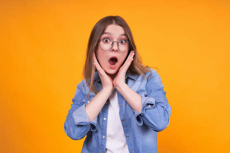 A stunned Caucasian young woman opens her eyes wide, suffocates in surprise, cannot believe the shocking news, is stunned, dressed in a denim shirt and round glasses.の写真素材