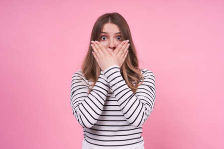An excited young lady model covers her mouth with her hands, not wanting to say something, a striped sweater with a pink background. The concept of not being heard of human feelings.Copy space.の写真素材