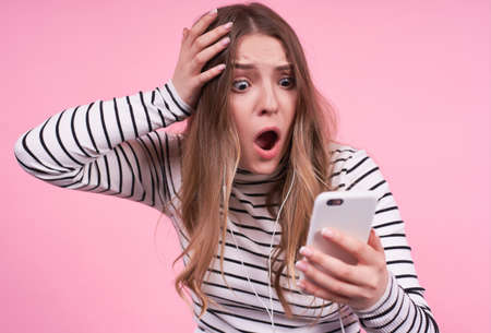 Young emotional Caucasian girl with a shocked surprised excited face with open mouth, clutching her head, looking at the phone, unhappy and not understanding what to do on a pink background.の写真素材
