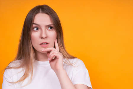 Close-up photo of an attractive charming Caucasian cute girl, touching her chin, meditating on something isolated with a yellow background, looking away with a doubtful and skeptical expression.の写真素材