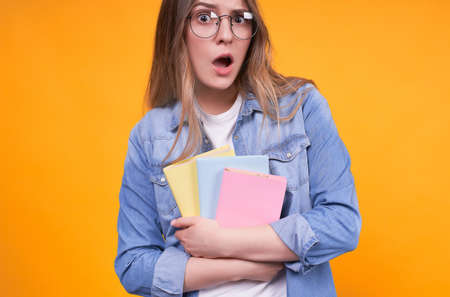 Young caucasian surprised amusing girl in glasses with notebooks, looking surprised and unexpected, with open mouth looking straight, thinking or making choices on a yellow background, copy space.の写真素材