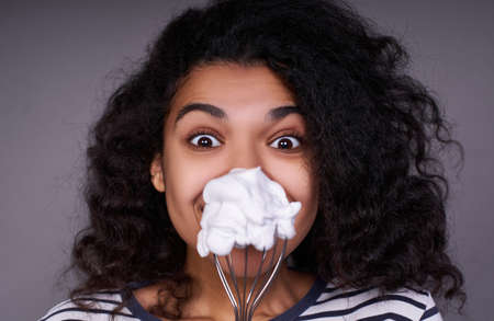 Photo of a dark-skinned girl, cooks at home, makes delicious cream, whips a white egg in a bowl, looks at the whisker with pleasure, prepares a mixture for baking. Culinary concept.の写真素材