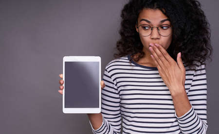 A shocked, amazed, amazed young dark-skinned woman in a striped sweater and glasses is holding a tablet in her right hand and covering her mouth with her left hand, she looks to the side.の写真素材