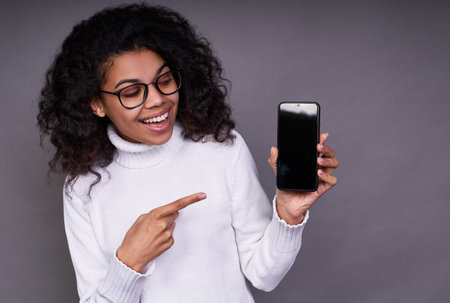 Close-up, a cheerful African-American girl, with curly hair, in a white sweater and glasses, holds a...