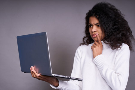Pensive african american girl isolated on gray studio background, hold a laptop. A girl of ethnic...
