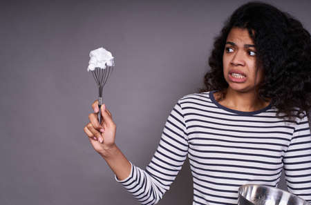 A young African-American girl holds a whisk with cream, looks with disgust, in her hands a bowl for cooking. He does not like to cook. Prepares a mixture for baking, cream for the cake.の写真素材