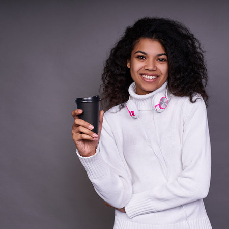 A vertical shot of a dreamy African-American girl, enjoys the taste of aromatic coffee, has lush...