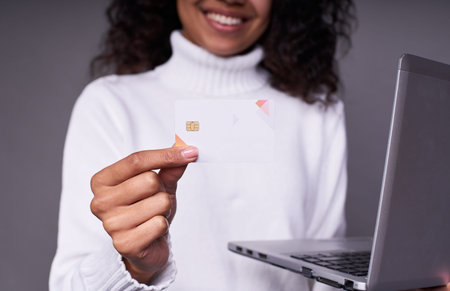 Close-up, smiling young dark-skinned girl in a white sweater, with a laptop, holds a white bank card...